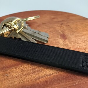 May include: A black key chain with a textured surface and a small logo on the end. The keychain is attached to a set of keys with a gold-coloured ring.