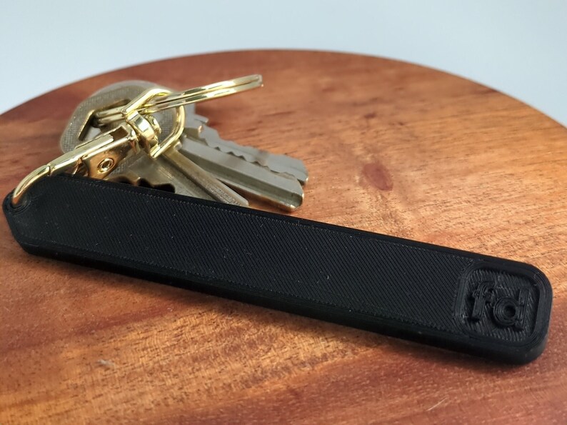 May include: A black key chain with a textured surface and a small logo on the end. The keychain is attached to a set of keys with a gold-coloured ring.
