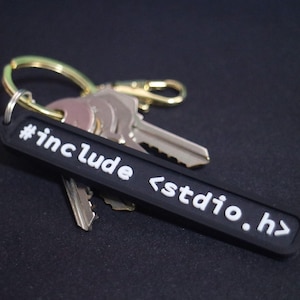 May include: Black keychain with white text that reads "#include <stdio.h>". The keychain is attached to a set of keys with a gold ring.