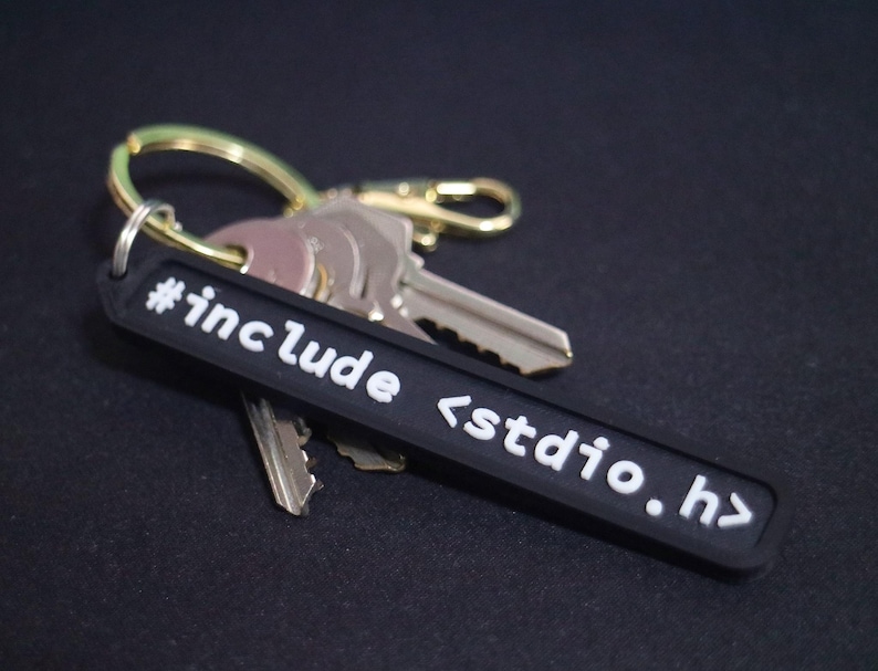 May include: Black keychain with white text that reads "#include <stdio.h>". The keychain is attached to a set of keys with a gold ring.