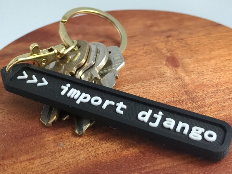 May include: Black keyring with white text that reads ">>> import django". The keyring is attached to a gold key ring with several keys.