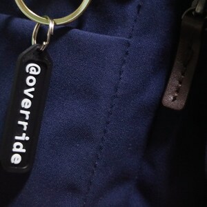 Puede incluir: Un llavero negro con el texto "@override" en letras blancas.