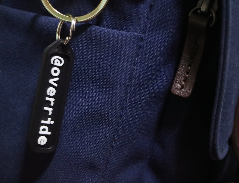 Puede incluir: Un llavero negro con el texto "@override" en letras blancas.