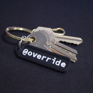 Puede incluir: Un llavero negro con el texto "@override" en letras blancas. El llavero est&aacute; unido a un juego de llaves plateadas.