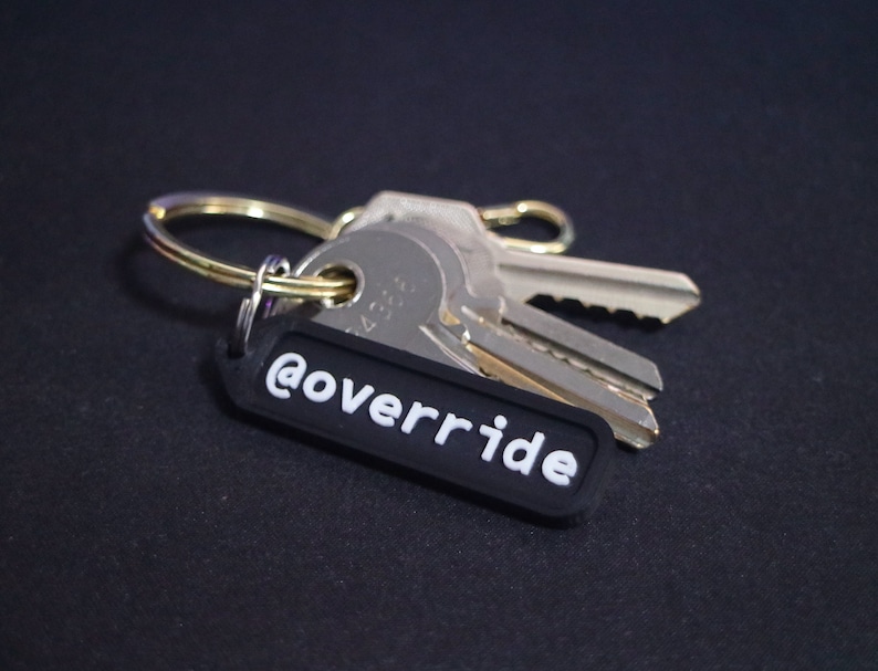 Puede incluir: Un llavero negro con el texto "@override" en letras blancas. El llavero est&aacute; unido a un juego de llaves plateadas.
