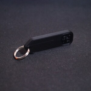 Puede incluir: Un llavero de pl&aacute;stico negro con un anillo de metal plateado. El llavero tiene un espacio rectangular en blanco para grabar.