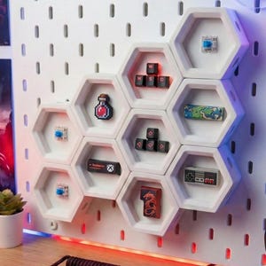 May include: White hexagonal wall shelves showcasing gaming accessories, such as keycaps, a pixelated potion, and a miniature controller. The shelves are mounted on a white pegboard. A small plant in a white pot is visible.