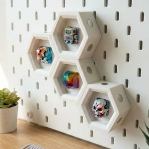 以下が含まれることがあります： 白い六角形の棚が白いペグボードに取り付けられており、それぞれ小さな装飾品を収納しています。アイテムには、ロボット、コイ、カラフルなクリスタル、スカルが含まれます。小さな鉢植えの多肉植物が木製の表面に置かれています。