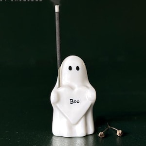 May include: A white ceramic ghost-shaped incense holder, 4.5 x 2.5 cm, with a heart that says "Boo." An incense stick is inserted, emitting smoke. The text "Incense Not Included" is also visible.
