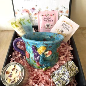 Può includere: Una confezione regalo contenente una tazza di ceramica blu con un disegno di fata, bustine di tè, tubetti di lozione per il corpo, una candela e un articolo a fantasia floreale. La tazza ha un manico viola ed è decorata con rose rosse.
