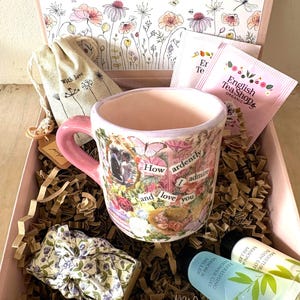 Könnte beinhalten: Eine Geschenkbox mit einer Keramiktasse mit rosa Henkel und einer Blumen-Collage mit dem Text How ardently I admire and love you. Enthalten sind ein kleiner Beutel mit Kordelzug, zwei English Tea Shop Teebeutel, ein Stoffbündel und zwei Tuben.