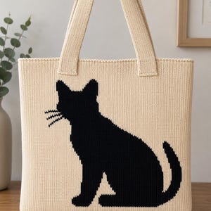 Könnte beinhalten: Eine cremefarbene Strick-Einkaufstasche mit einer großen, schwarzen, massiven Silhouette einer sitzenden Katze auf der Vorderseite. Die Tasche hat zwei passende cremefarbene Strick-Schulterriemen und eine rechteckige Form mit einem sichtbaren Strickmuster.