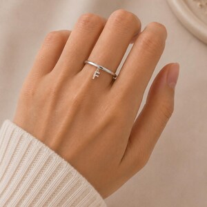 Può includere: Un anello color argento con una fascia sottile e liscia, caratterizzato da un piccolo ciondolo pendente a forma di lettera F. Il ciondolo a forma di lettera è impreziosito da minuscole pietre trasparenti e scintillanti, per un aspetto delicato.