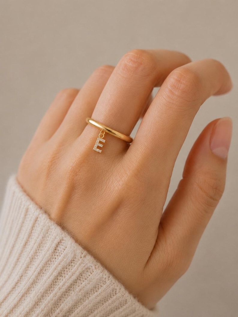 May include: A gold-toned ring featuring a smooth, polished band and a small, dangling charm in the shape of the letter E. The letter charm is encrusted with tiny, clear, sparkling stones that catch the light, creating a delicate and refined appearance.