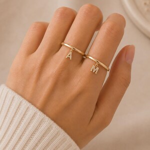 May include: Two thin gold rings, each featuring a small, dangling letter charm. One ring displays the letter A and the other displays the letter M. Both letter charms are encrusted with tiny, clear, sparkling stones along their surfaces.