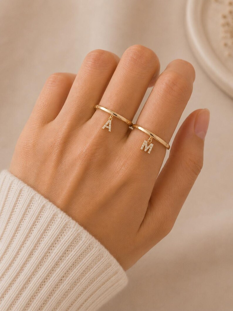 May include: Two thin gold rings, each featuring a small, dangling letter charm. One ring displays the letter A and the other displays the letter M. Both letter charms are encrusted with tiny, clear, sparkling stones along their surfaces.