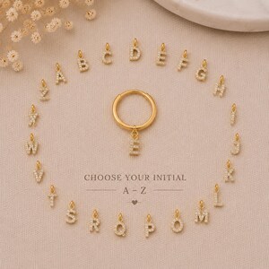 May include: A gold hoop earring featuring a removable letter E charm, surrounded by a collection of individual gold letter charms from A to Z, each encrusted with small clear stones. The text CHOOSE YOUR INITIAL A-Z is printed in the center.