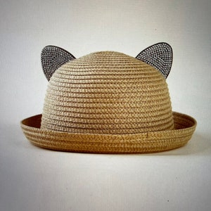 May include: A straw hat with a wide brim and cat ears adorned with rhinestones. The hat is a light brown color.