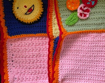 May include: A colorful crocheted blanket with squares in pink, blue, green, and white. The squares are bordered with orange and red yarn. One square features a crocheted yellow face with a smile.