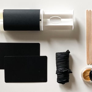 Op de afbeelding: Een set zwarte en witte knutselbenodigdheden voor het maken van kaarsen. De set bevat een zwarte kaarsenmaakset, twee zwarte kaarten, een zwarte plastic zak, een potje gouden glitter, een set houten stokjes en een zwart deksel.