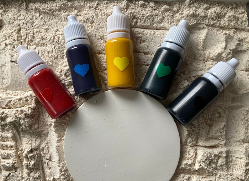 K&ouml;nnte beinhalten: F&uuml;nf kleine Flaschen mit fl&uuml;ssiger Farbe und wei&szlig;en Verschl&uuml;ssen. Die Flaschen sind in einem Halbkreis auf einer wei&szlig;en Oberfl&auml;che angeordnet. Die Farben der Farbe sind rot, blau, gelb, gr&uuml;n und schwarz.