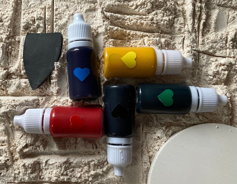 K&ouml;nnte beinhalten: F&uuml;nf kleine Flaschen mit Farbe in verschiedenen Farben und wei&szlig;en Verschl&uuml;ssen. Jede Flasche hat einen kleinen herzf&ouml;rmigen Aufkleber in einer anderen Farbe. Die Farben sind blau, gelb, rot, gr&uuml;n und schwarz.