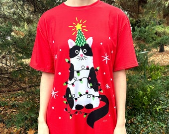 May include: Red t-shirt dress with a black and white cat wearing a Christmas tree hat and wrapped in Christmas lights.