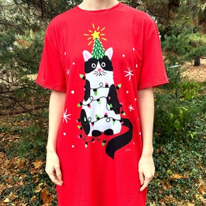 May include: Red t-shirt dress with a black and white cat wearing a Christmas tree hat and wrapped in Christmas lights.