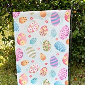 May include: A blue and white garden flag with a pattern of colorful Easter eggs. The eggs are decorated with flowers, stripes, and polka dots.