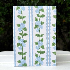 May include: A rectangular greeting card featuring a floral pattern. The design includes vertical stripes of light blue with a scalloped edge, and white space. The card is decorated with illustrations of blue hydrangea flowers and green leaves.