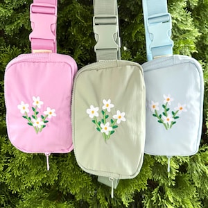 May include: Three rectangular belt bags in pastel pink, sage green, and light blue. Each bag features an adjustable strap with a plastic buckle and an embroidered design of white flowers with green stems and leaves on the front zippered compartment.