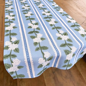 May include: A blue table runner with a white floral pattern and white scalloped edges. The pattern features white flowers with green leaves on a light blue background.