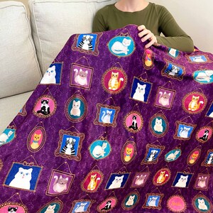 May include: Purple fleece blanket with a repeating pattern of cats in gold frames. The cats are in various poses and colors, including black, white, orange, and gray.