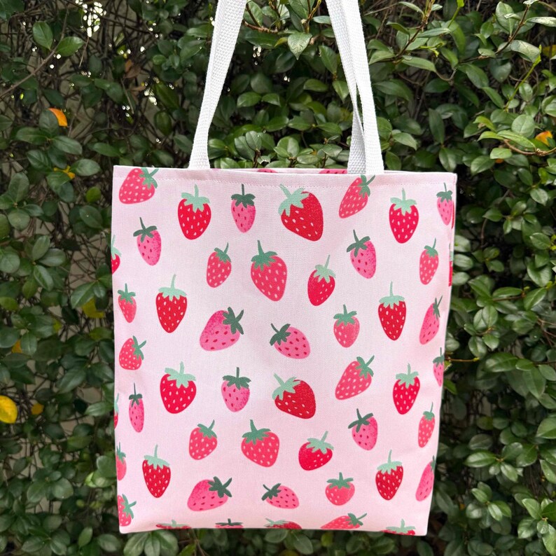 May include: A pink canvas tote bag with a white handle and a repeating pattern of red strawberries on a pink background.