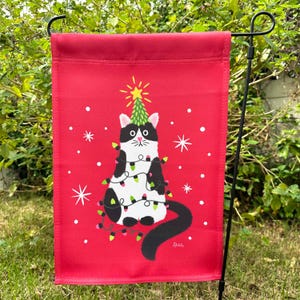 May include: A red Christmas garden flag with a black and white cat wearing a string of lights and sitting on top of a green Christmas tree. The flag has a white snowflake pattern and the text "Zibbit" in the bottom right corner.