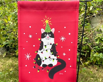 May include: A red Christmas garden flag with a black and white cat wearing a string of lights and sitting on top of a green Christmas tree. The flag has a white snowflake pattern and the text "Zibbit" in the bottom right corner.
