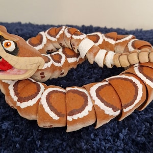 May include: A brown and white segmented rattlesnake toy with a wide open mouth and red tongue. The toy is made of wood and painted to look like a real rattlesnake.