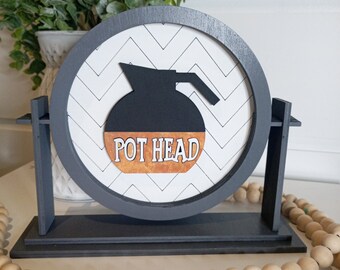May include: A round wooden sign with a black and white chevron pattern. The sign features a black silhouette of a coffee pot with the text "POT HEAD" in white on a brown background.