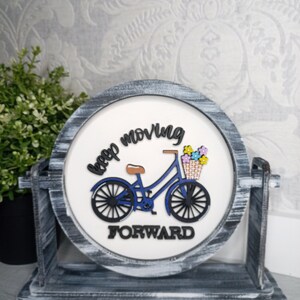 May include: A round wooden sign with a distressed gray finish. The sign features a blue bicycle with a basket of flowers and the text "keep moving forward".
