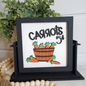 Peut inclure: Une pancarte encadrée de noir avec un fond blanc et le texte "CARROTS 5¢" en lettres noires. Un seau en bois brun avec des fanes de carottes vertes et des carottes orange est représenté sous le texte.