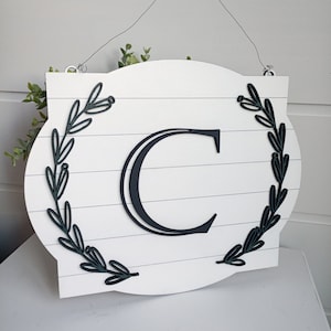 Könnte beinhalten: Ein weißes Holzschild mit einem schwarzen Buchstaben C in der Mitte. Das Schild ist mit schwarzen und grünen Blattwerk verziert, das sich auf beiden Seiten des Buchstabens befindet. Das Schild hat einen Drahtbügel.