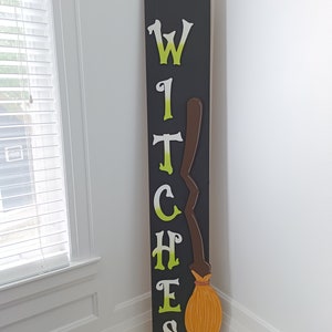 Op de afbeelding: Een zwart houten bord met het woord "welcome" in witte verf. Het bord heeft ook het woord "witches" in groene en witte verf geschreven met een bruine en oranje bezem rechts van het woord.
