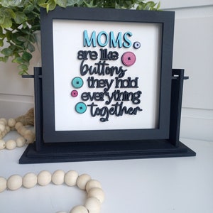 Puede incluir: Un letrero enmarcado en negro con las palabras "MOMS are like buttons they hold everything together" en azul, rosa y verde. El letrero está sobre un fondo blanco. Una guirnalda de cuentas de madera está en primer plano.