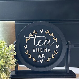 May include: Black round sign with a gold floral design and the text "tea a hug in a mug". The sign is on a black stand.