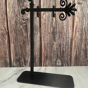 May include: Black metal earring display stand with a rectangular base and a decorative flourish. The stand has a vertical post and a horizontal bar with multiple earring holes. The background is a wooden wall.
