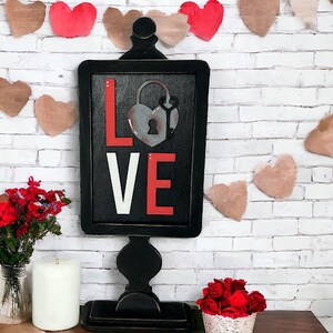 May include: Black wooden sign with the word "LOVE" in white and red letters. A rusty padlock with a key is in the centre of the sign. The sign is on a black stand with a decorative top. The sign is surrounded by a garland of brown and red paper hearts.