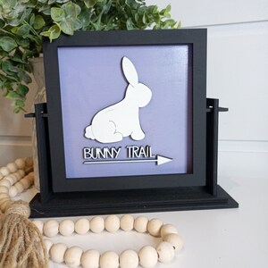 Puede incluir: Un letrero enmarcado en negro con una silueta de conejo blanco y el texto "Bunny Trail" con una flecha apuntando hacia la derecha. El letrero está sobre una superficie blanca con una guirnalda de cuentas de madera.