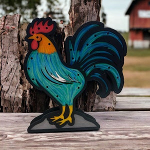 Puede incluir: Una figura de gallo de madera con un cuerpo pintado de azul y verde, una cresta roja y una base pintada de negro. El gallo está de pie sobre una superficie de madera.