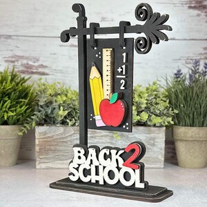 May include: Black wooden sign with the words "Back 2 School" in white letters. The sign also features a black chalkboard with a yellow pencil, a red apple, and a ruler with the equation "1 + 1/2".
