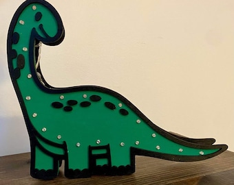 May include: A green dinosaur-shaped night light with black outlines and white lights. The dinosaur is standing on a wooden surface.